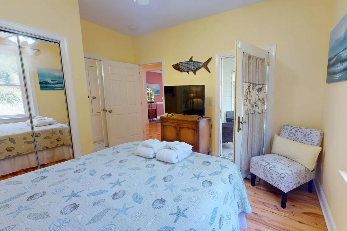 A cozy bedroom with a decorative quilt on the bed, wall-mounted TV unit, armchair for reading, and a fish-themed decoration that adds a coastal touch. Ample natural light pours in, enhancing the room's inviting ambiance.