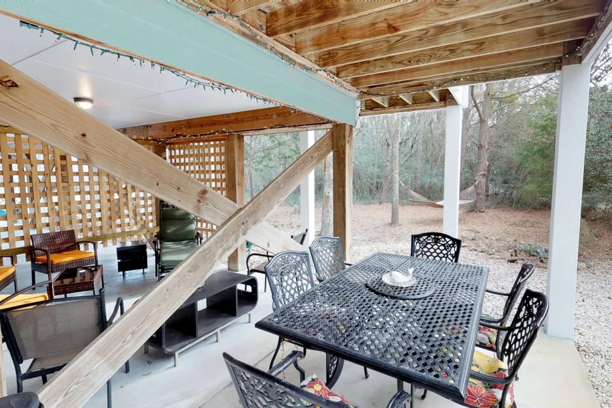 A welcoming outdoor deck space with a large wooden dining table under wooden beams, a lattice wall creating privacy, and a hammock strung in the background. The area is surrounded by greenery and bathed in natural light, creating a relaxed environment for dining or lounging.