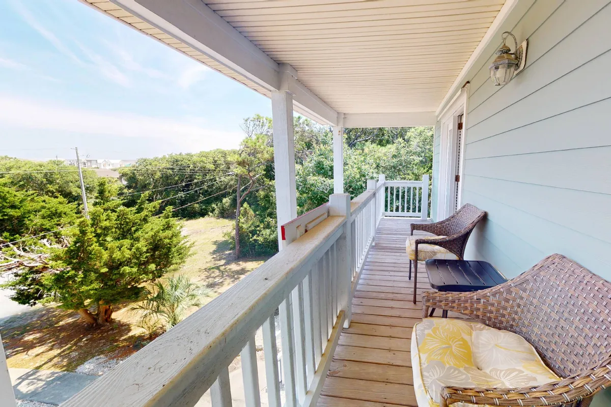 A wooden deck with classic wicker chairs overlooking lush greenery, framed by a sturdy wooden railing. The scene exudes tranquility, offering a spot to enjoy morning coffee or simply take in the peaceful, natural surroundings.