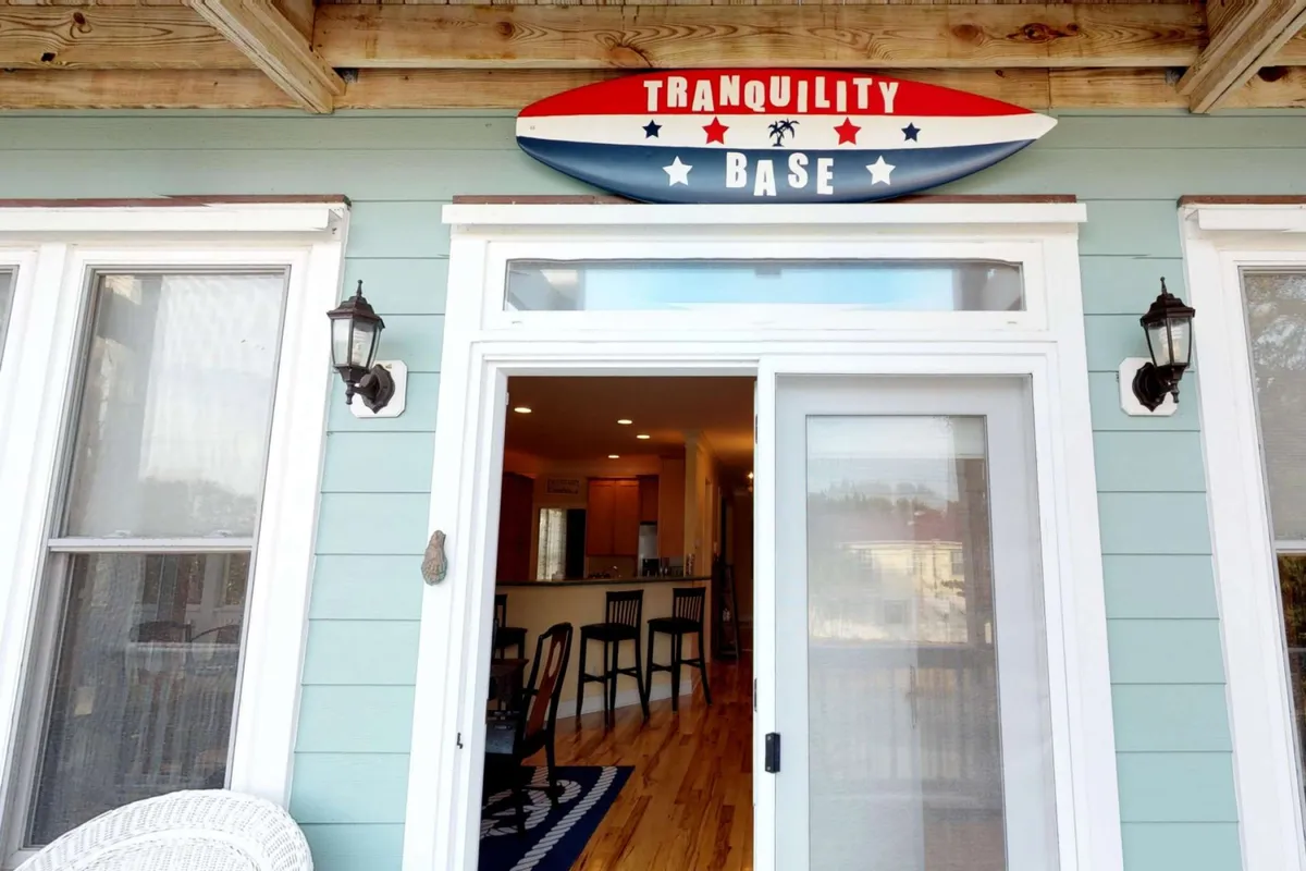 An inviting exterior porch scene featuring a sign with 'TRANQUILITY BASE,' a white wicker chair, and a view into the kitchen area through the window. The porch suggests a seamless transition from indoor culinary spaces to outdoor relaxation, hinting at the experience of preparing meals and then enjoying them outside.