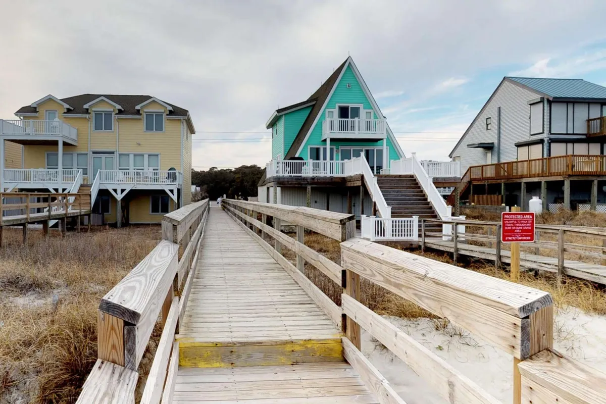 A boardwalk scene leads towards colorful beach houses, surrounded by natural coastal landscaping. The image captures the essence of beachside exploration, evoking a sense of casual adventure and carefree play. It's easy to imagine guests strolling with their dogs along the boardwalk toward the sandy beach.