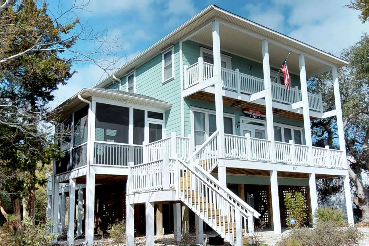 The hero image showcases the striking bright blue exterior of a spacious single-family beach house in Kure Beach, North Carolina. A large, inviting porch wraps around the front, adorned with comfortable seating for gatherings and relaxation. An American flag gently waves, adding a sense of warmth and welcome. The setting suggests opportunities for sunset views from the balcony, sipping morning coffee on the porch, and hosting evening barbecues with family and friends.