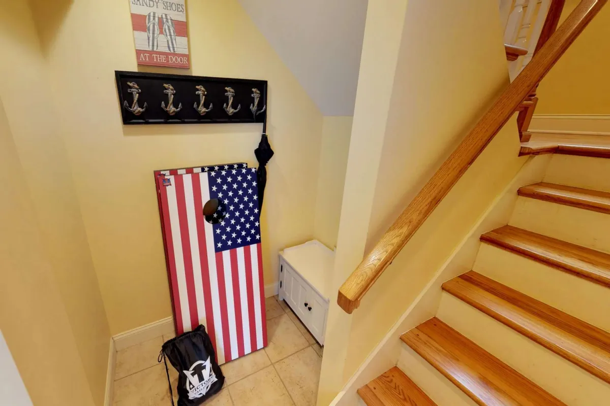 A casual indoor-outdoor transition area featuring an American flag-themed cornhole board by a wooden handrail, anchor-themed wall hooks, and a storage bench. The space feels playful and family-friendly, ready for games and welcoming guests.