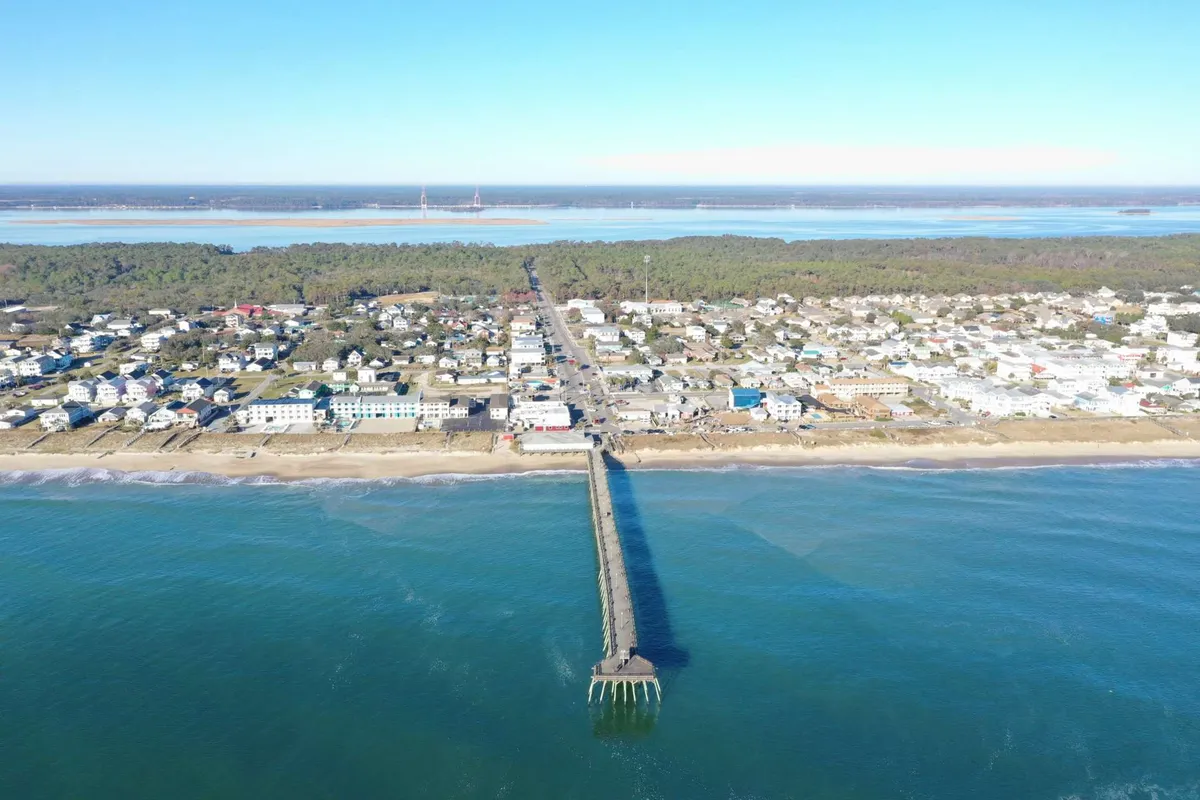 A sweeping view of the Kure Beach Pier extending over the ocean, with the beach and town in the background. The pier is bustling with activity, inviting visitors to fish, take a stroll, or enjoy the local scene. The ocean glimmers under soft light, while the town’s charm adds to the lively ambiance.