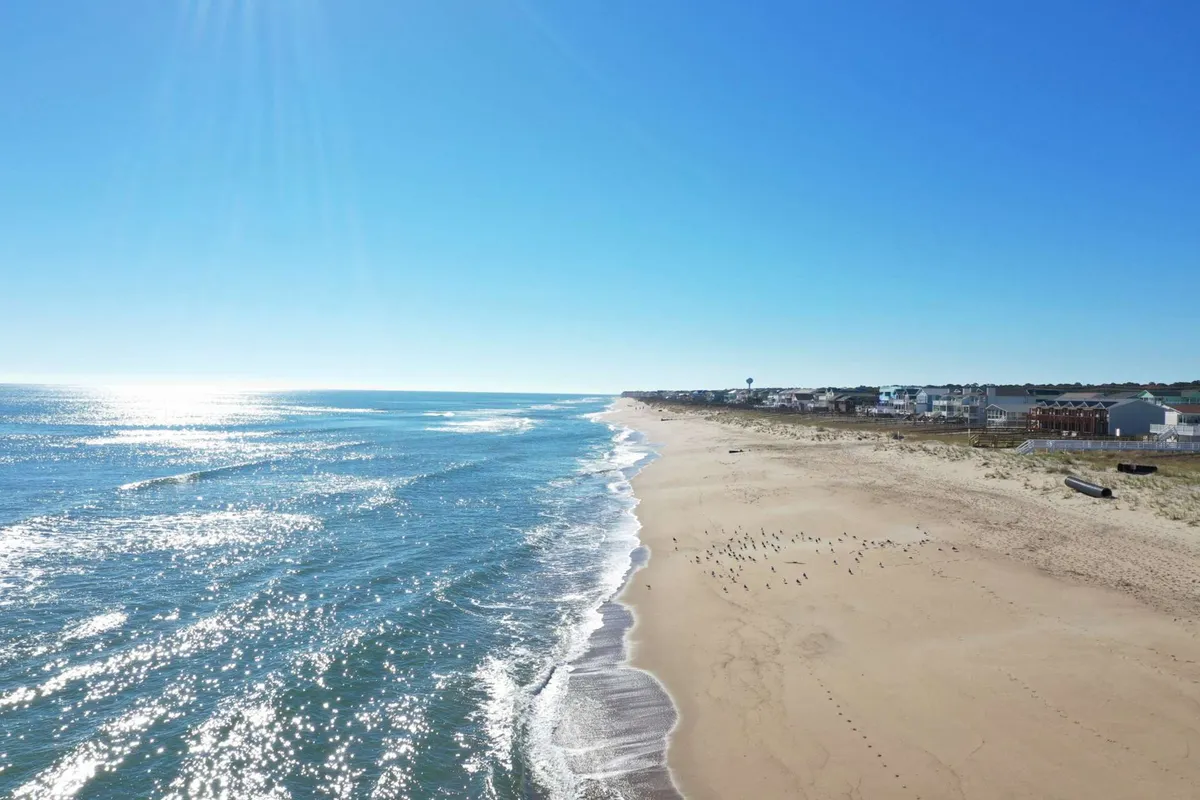 A wide-angle view of Kure Beach with calm ocean waves lapping the golden sandy shore. The expansive beach stretches into the distance, dotted with birds and bordered by a tranquil shoreline. The mood is serene, conjuring images of leisurely walks, beachcombing, and peaceful sunbathing.