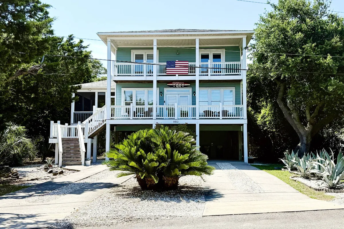 The exterior of Tranquility Base, framed by balconies and a palm tree, with an inviting driveway leading up to the house. The scene suggests easy access to and from the beach, and the house’s spacious decks are visible—perfect for midday siestas and enjoying ocean breezes between outings.