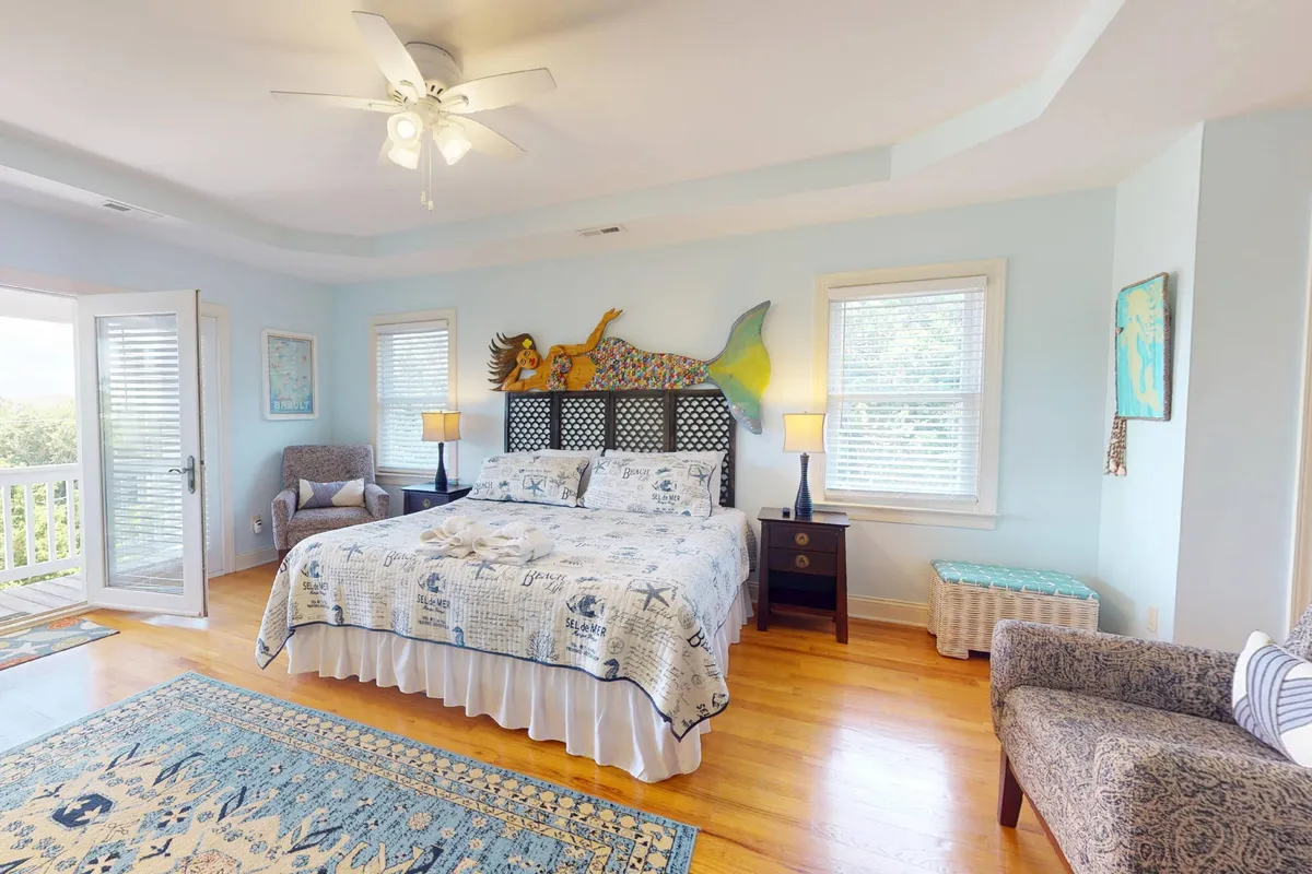 A bright and whimsical bedroom with a bed featuring a mermaid headboard, a decorative quilt, large sunlit windows, and a comfy armchair. The playful decor appeals to children and families, offering a retreat for rest or imagination.