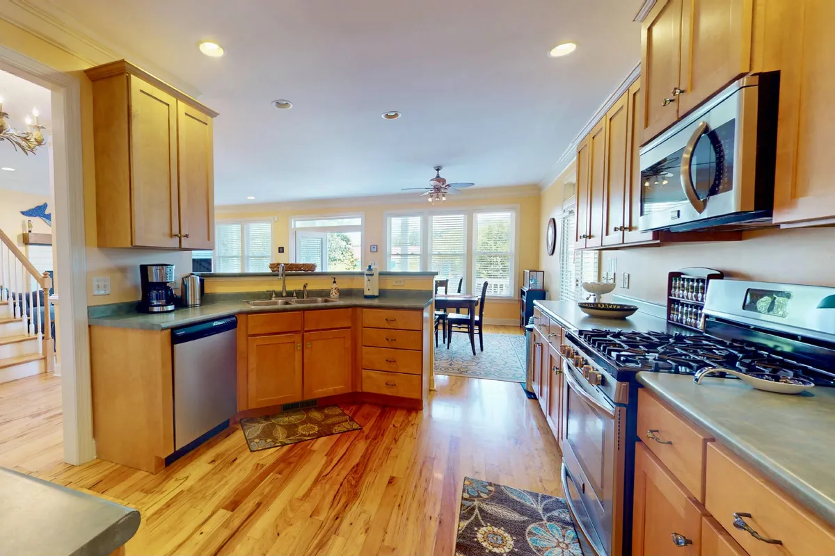 This image highlights the kitchen island with stainless steel appliances, natural light streaming in from windows, and a view into a dining area. The scene is styled for welcoming family gatherings and culinary activities, showcasing both the kitchen and its seamless connection to the dining space.