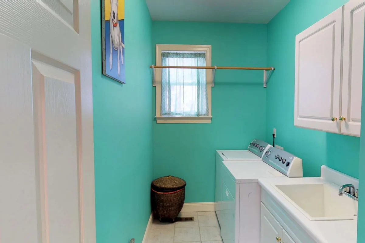 A playful laundry room with turquoise walls, a modern washer and dryer, a laundry sink, and a decorative dog painting. A laundry basket is neatly placed nearby, suggesting ample storage and functionality. The cheerful decor is both family- and pet-friendly.