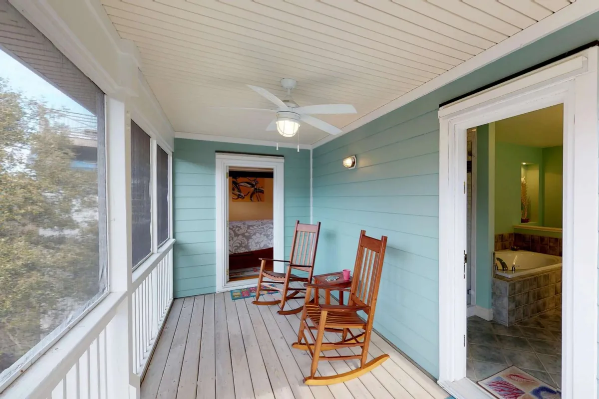 A broad exterior view of the home's screened porch, featuring inviting rocking chairs and direct access to interior rooms. The space is sunlit and open, seamlessly connecting the indoors with the outdoors; ideal for relaxing with pets after a beach adventure. The porch overlooks sunrise views and is perfect for sharing breezy afternoons or unwinding after a day on the sand.
