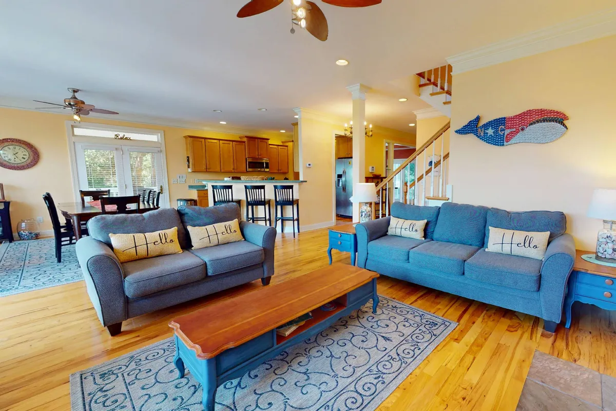 A spacious, inviting living area featuring two blue sofas, a wooden coffee table, vibrant wall art, and an open kitchen in the background. The room is filled with natural light and designed for gatherings, with comfortable seating and a layout that encourages socializing.
